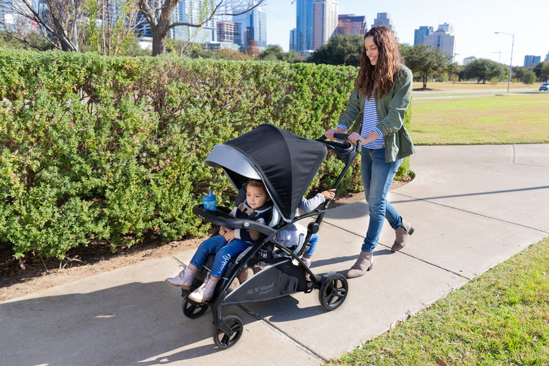 Mom and her two children strolling outdoor in the park with the Baby Trend Sit N' Stand Shopper Stroller.